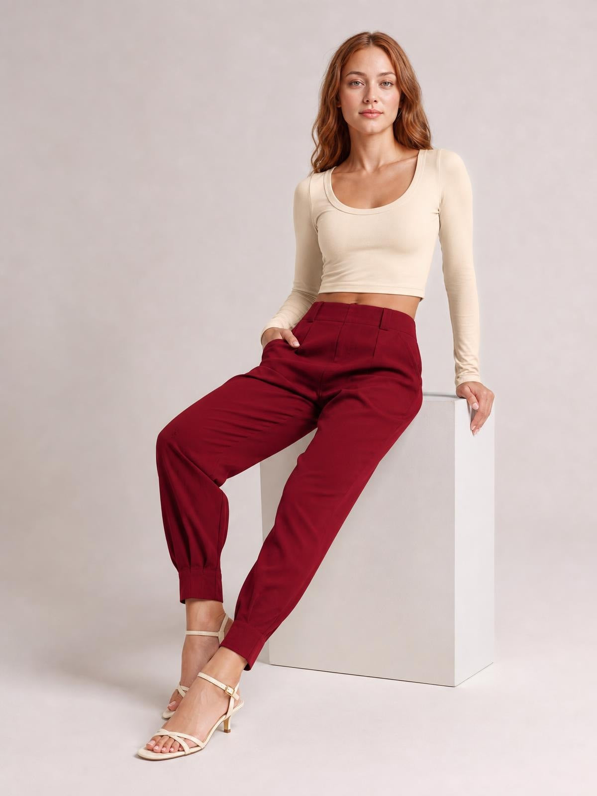 Model seated in high-waist cropped pants in deep burgundy, featuring a tapered silhouette, button closure, belt loops, and side pockets.