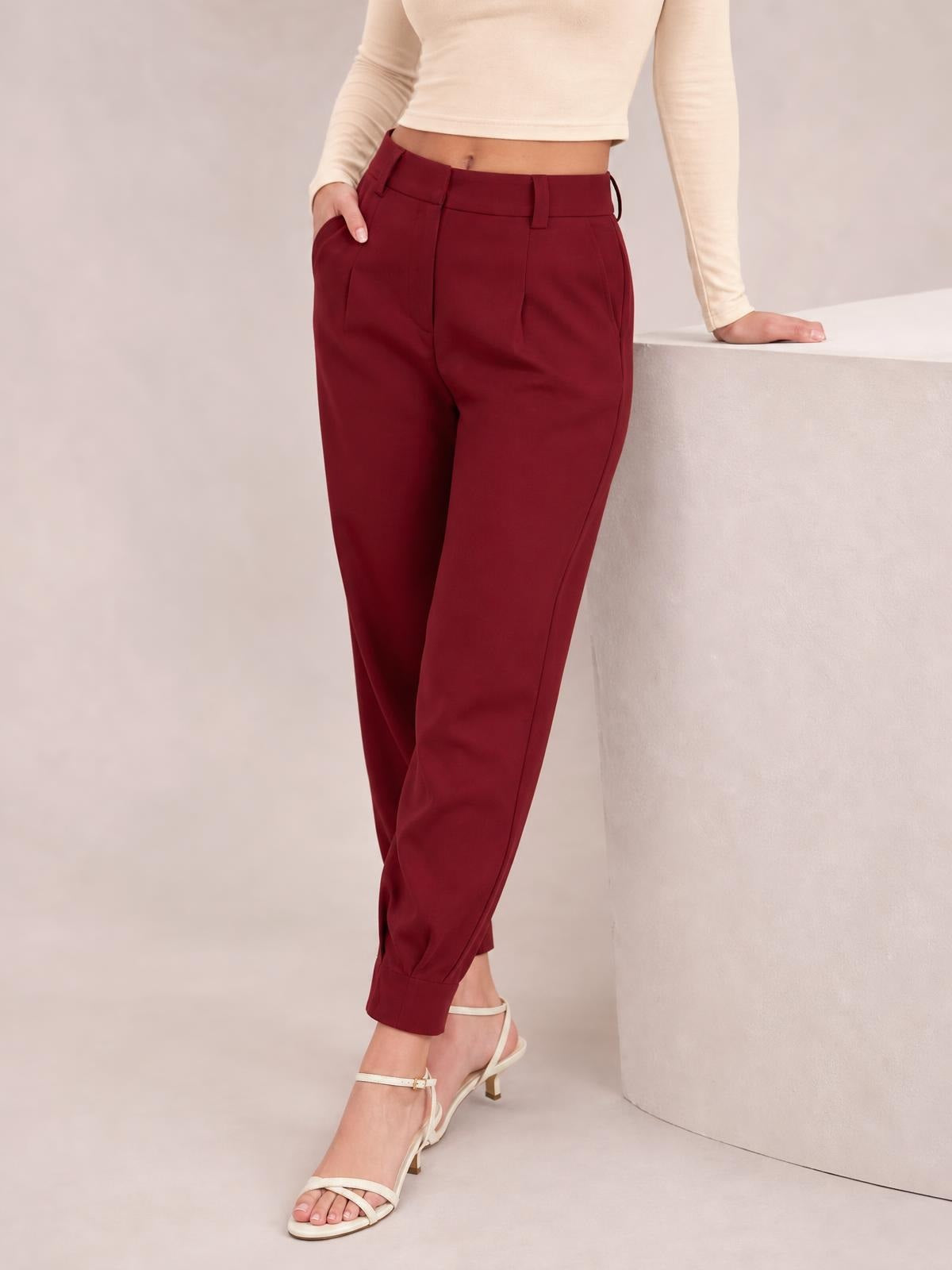 A model wears burgundy high waist cropped pants with a tapered silhouette, belt loops, side pockets, and cuffed hems, posing with one hand in a pocket.