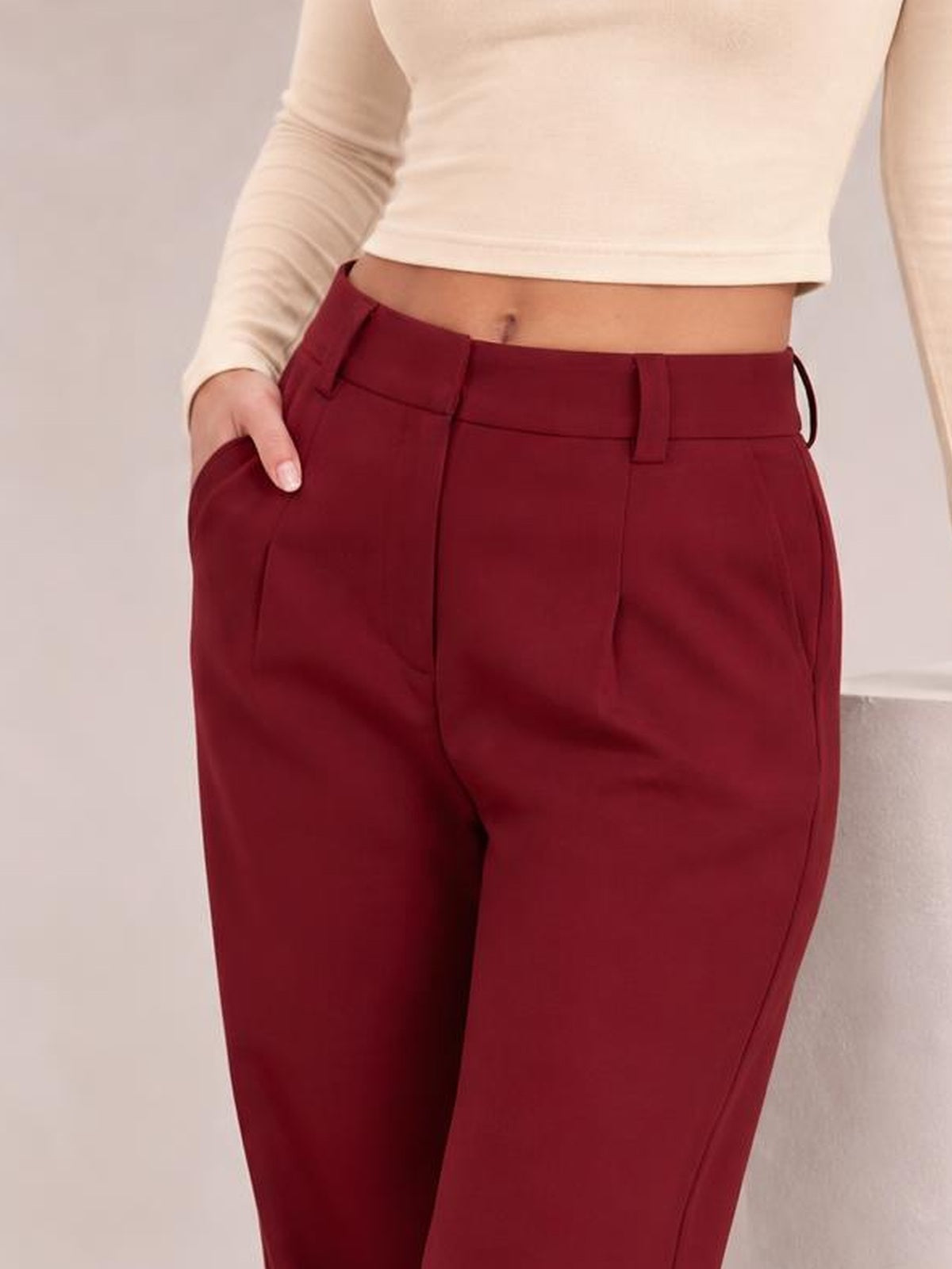 A model wears burgundy high waist cropped pants with a structured waistband, belt loops, front pleats, and side pockets, posing with one hand tucked inside.