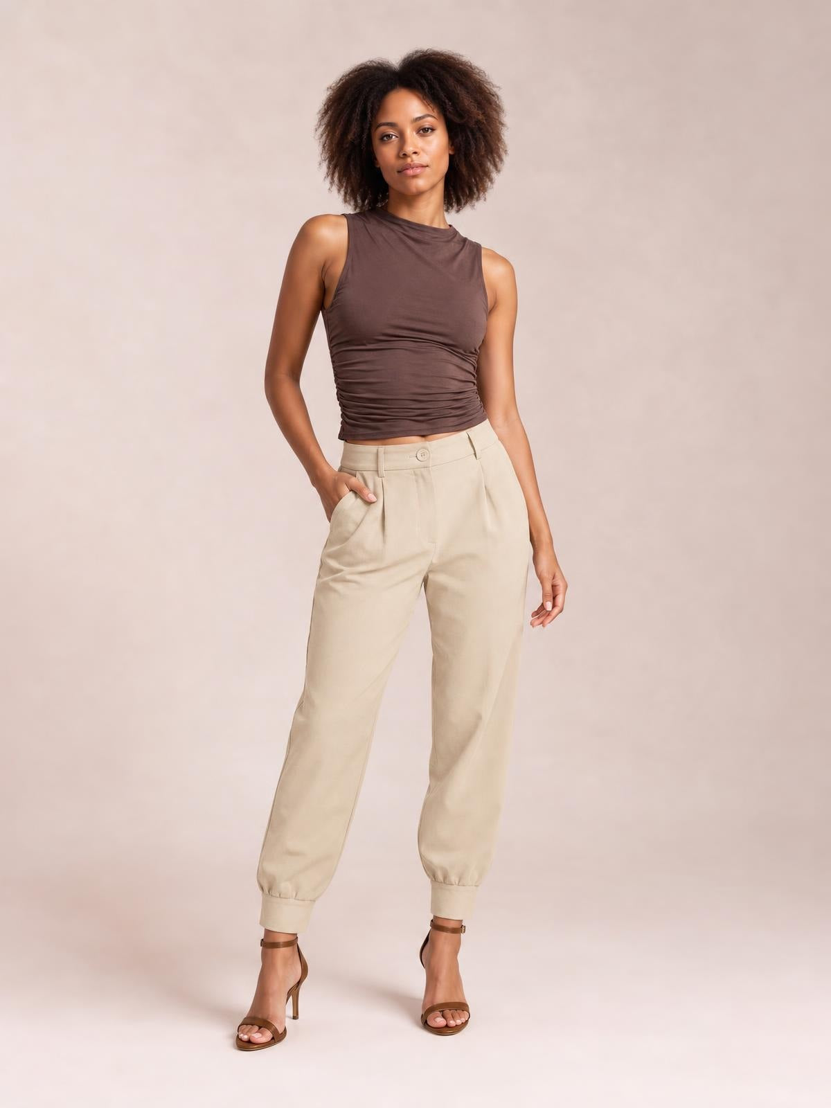 A model poses in high-waist cropped pants in beige, featuring a buttoned front, side pockets, relaxed tapered legs, and gathered ankle cuffs.
