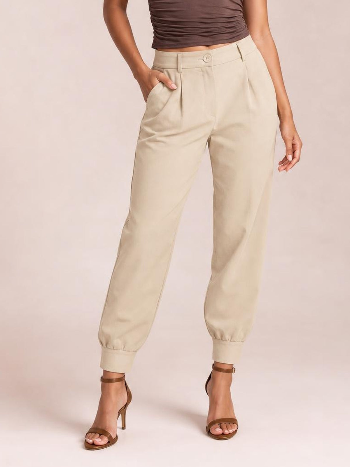 A model wears beige high-waist cropped pants with a buttoned waistband, side pockets, relaxed tapered legs, and cuffed hems, resting one hand in a pocket.