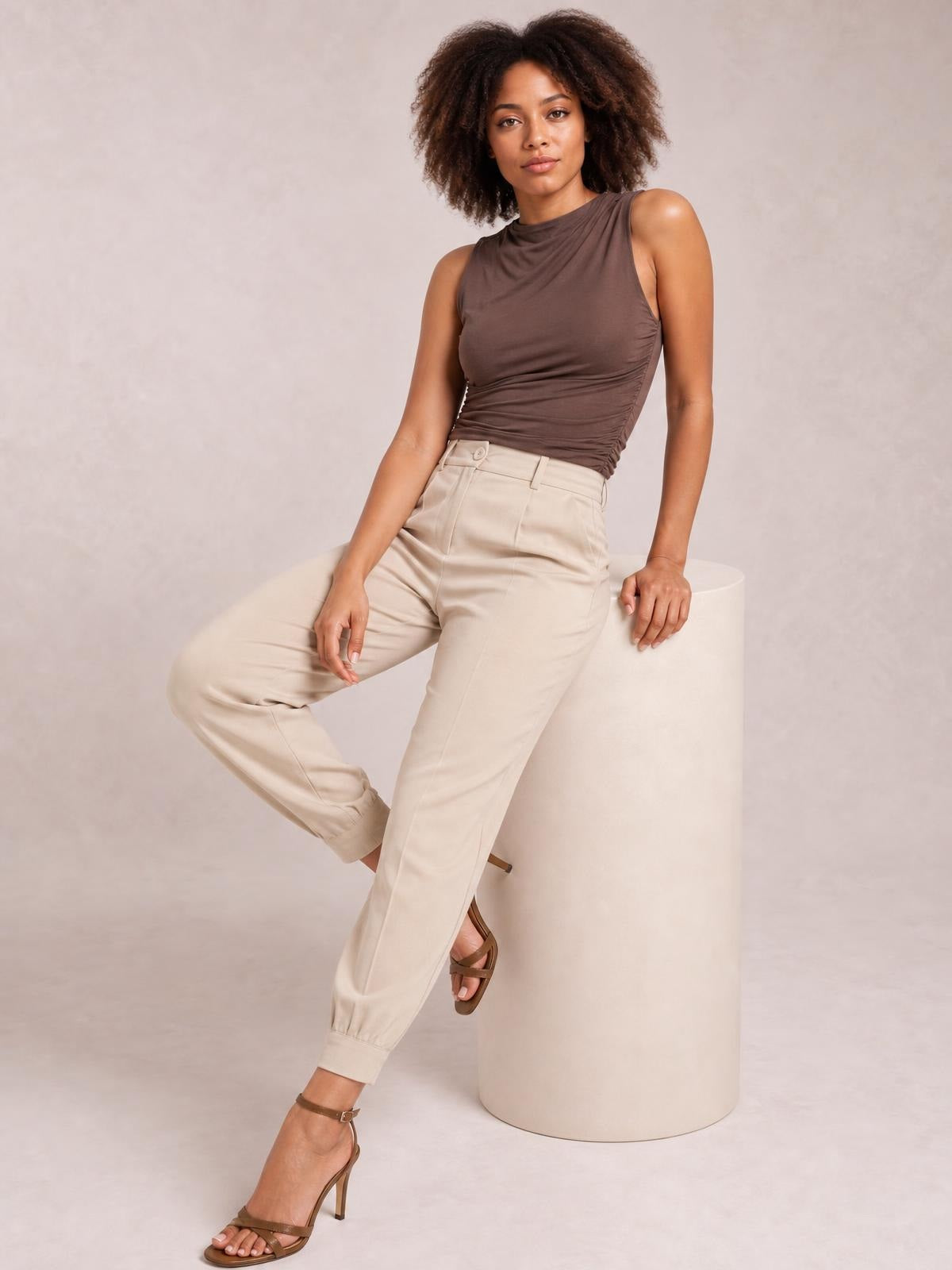 Model poses in beige high-waist cropped pants with a buttoned waistband, belt loops, front pockets, tapered legs, and cuffed hems.