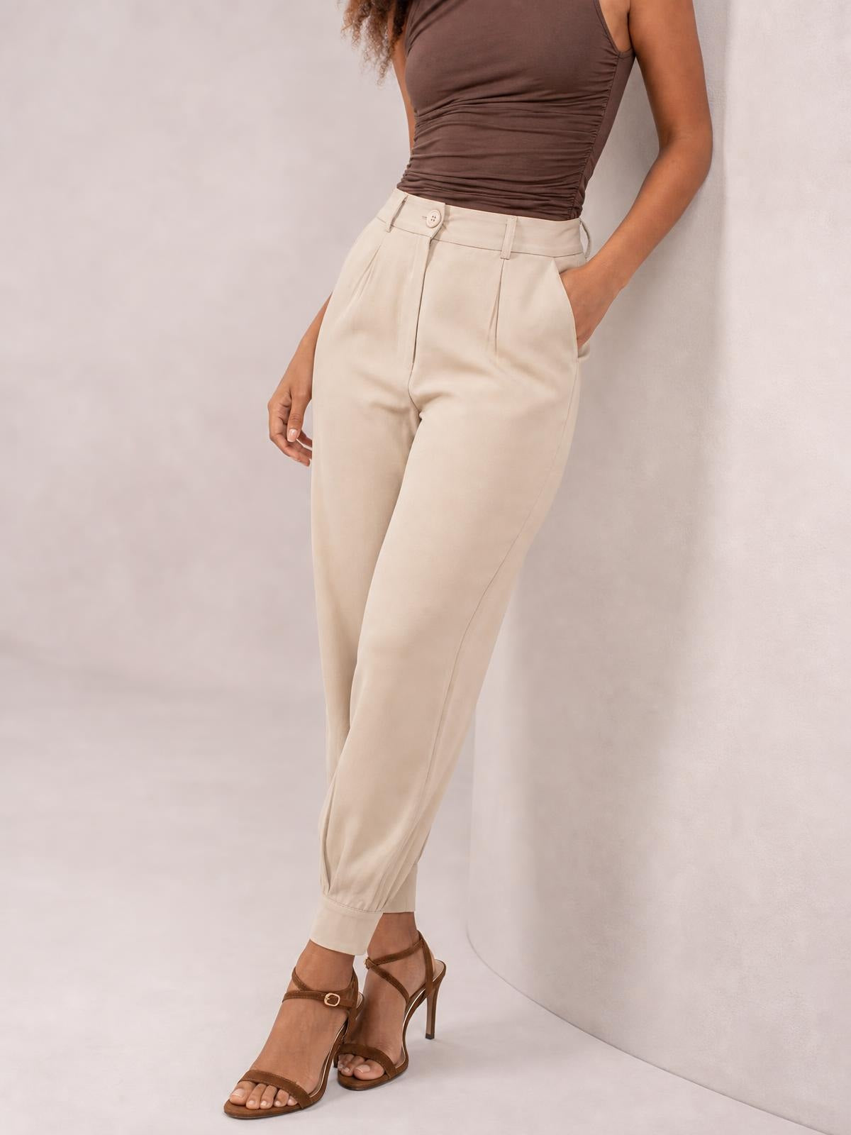 A model poses in cream high waist cropped pants with a buttoned waistband, belt loops, front pleats, side pockets, and tapered ankle cuffs.