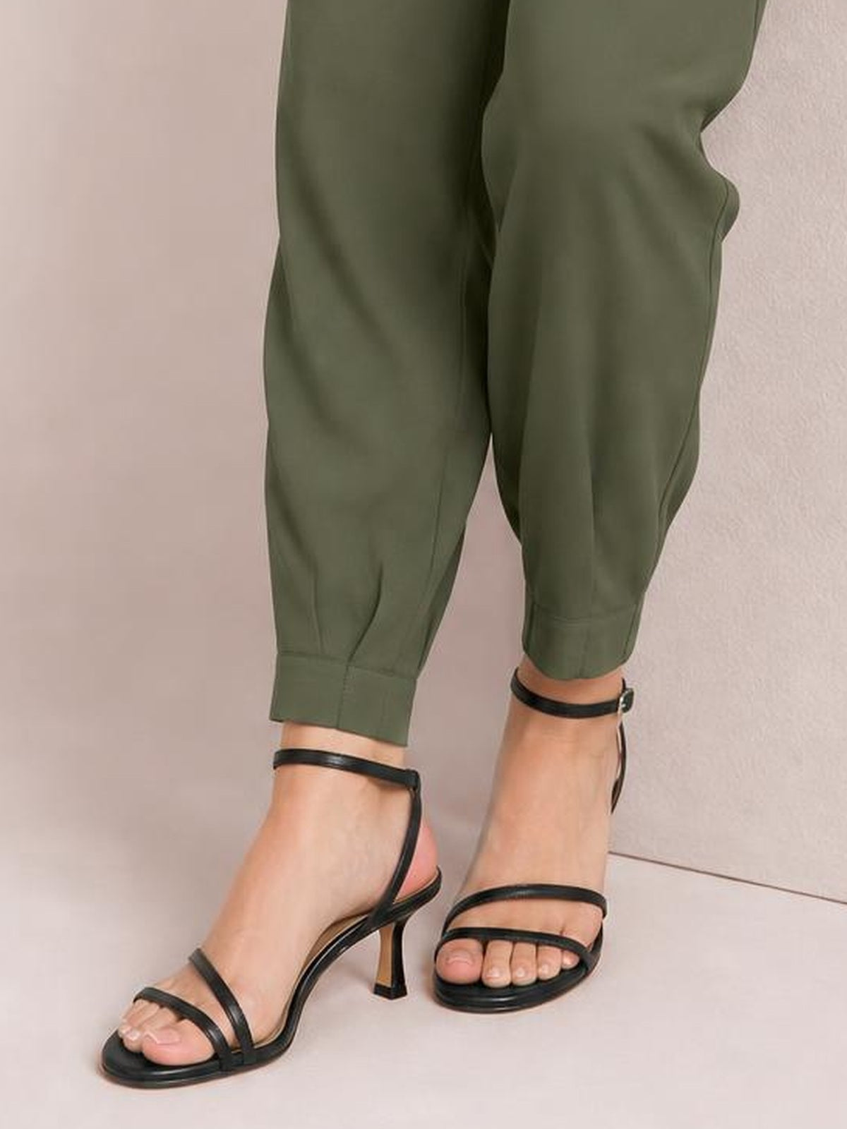 Model wears olive green high-waist cropped pants with a tapered silhouette and neat ankle cuffs, styled with black strappy heels.