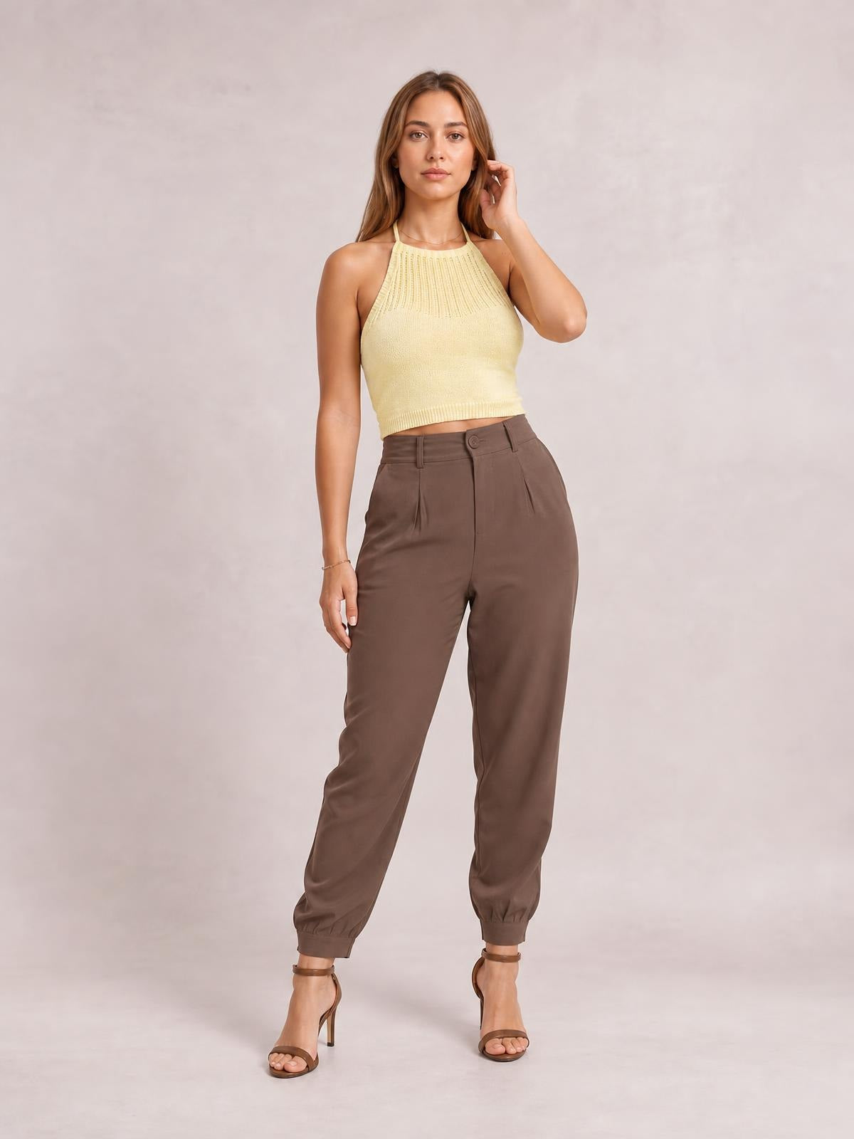 A model poses in brown high-waist cropped pants with a buttoned front, belt loops, pleated construction, side pockets, and a tapered ankle-length silhouette.