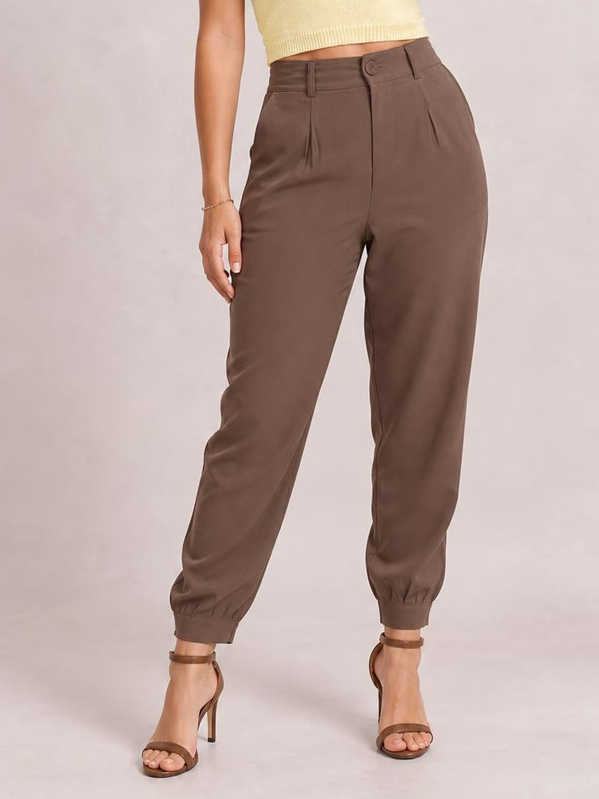 A model stands with one hand at her side, wearing brown high-waisted cropped pants with a buttoned waistband, front pleats, pockets, and tapered elastic cuffs.