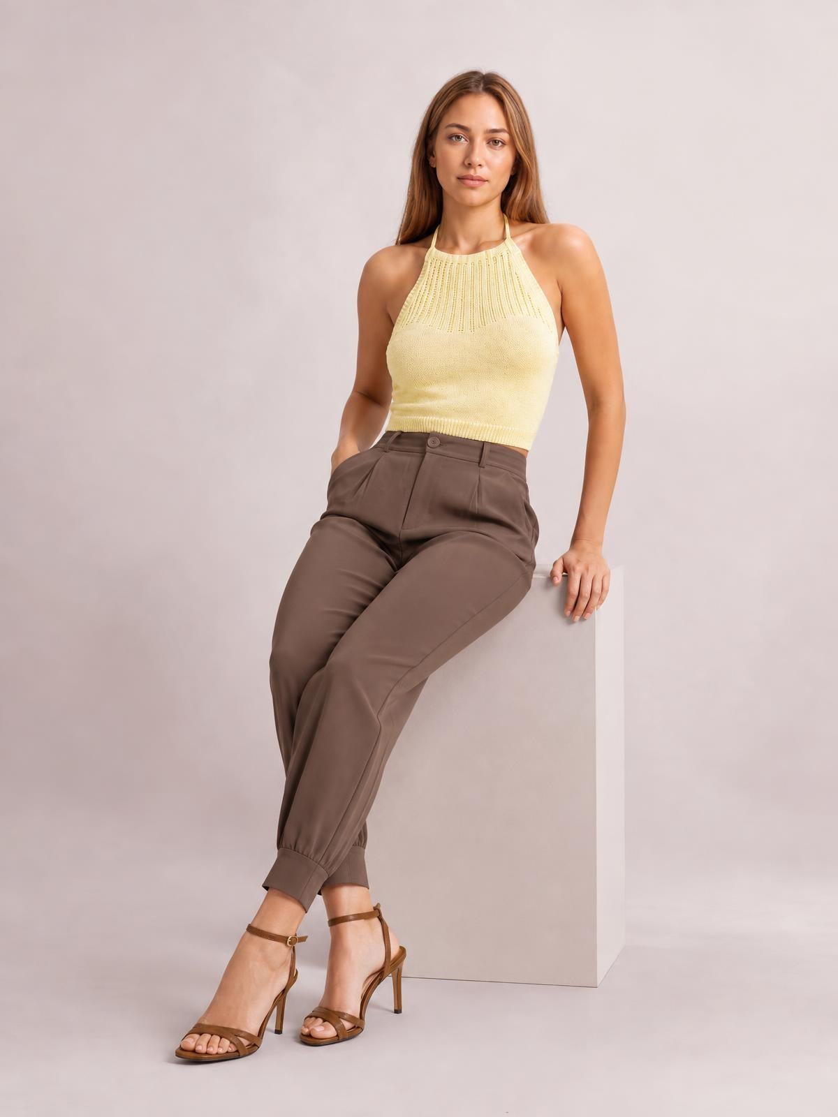 A model sits in brown high-waisted cropped pants with a button closure, slanted pockets, tapered legs, and cuffed ankle hems.