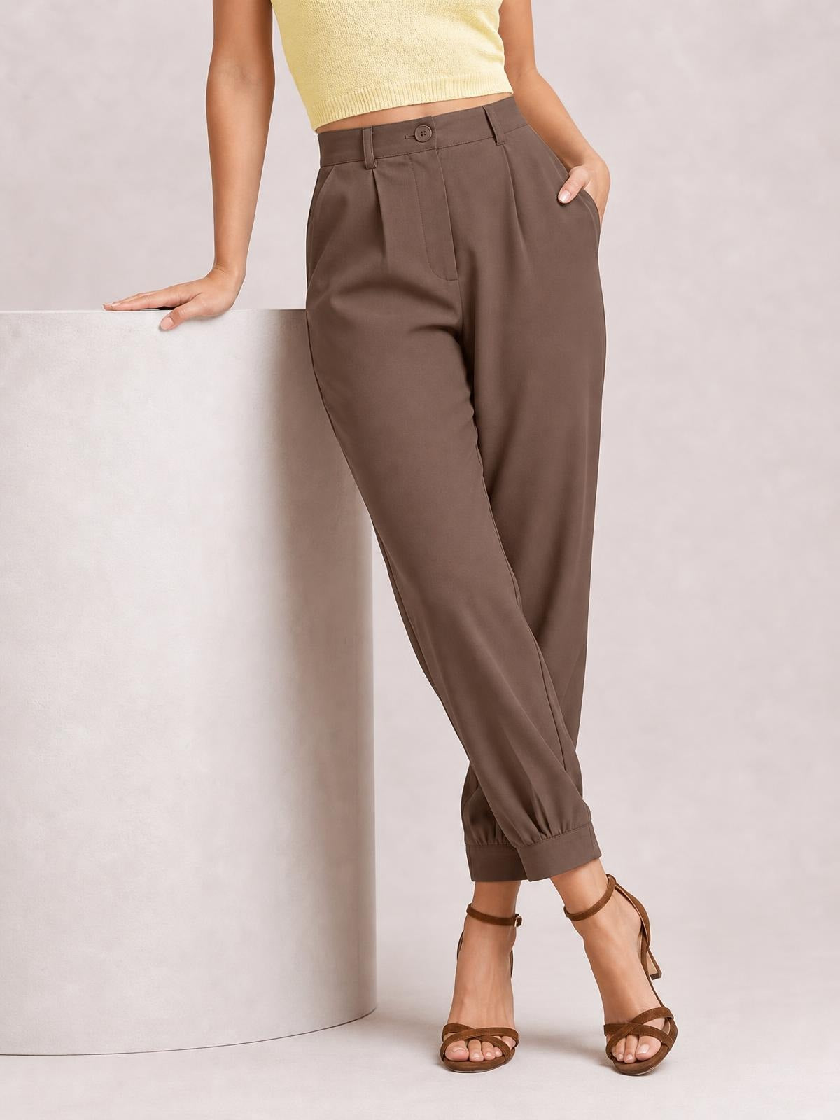 A model poses with one hand in a pocket, wearing brown high-waisted cropped pants with a buttoned waistband, belt loops, tapered legs, and cuffed hems.