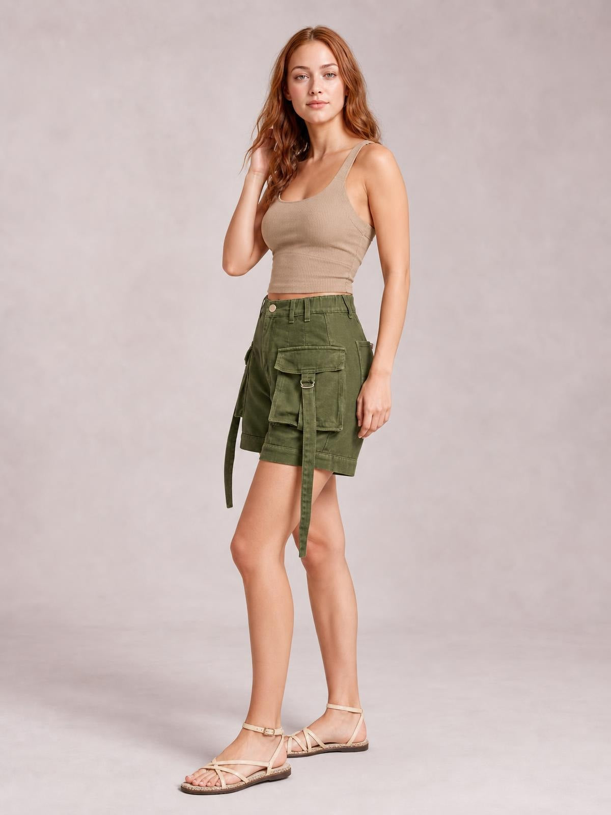 Model poses in olive green high-waist cargo shorts with a relaxed silhouette, belt loops, stitched panels, and multiple flap pockets with hanging straps.