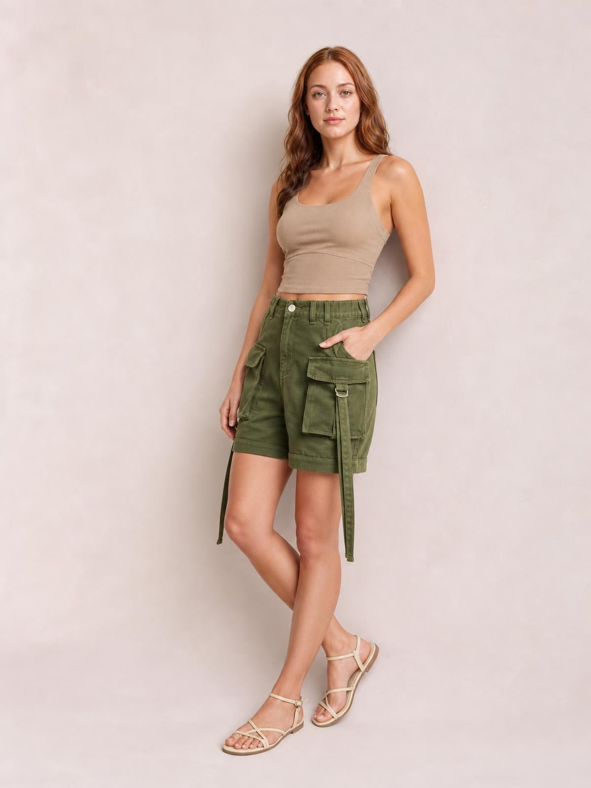 Model wearing olive high-waist cargo shorts with a relaxed silhouette, belt loops, button closure, and multiple flap pockets, posing with one hand in a pocket.