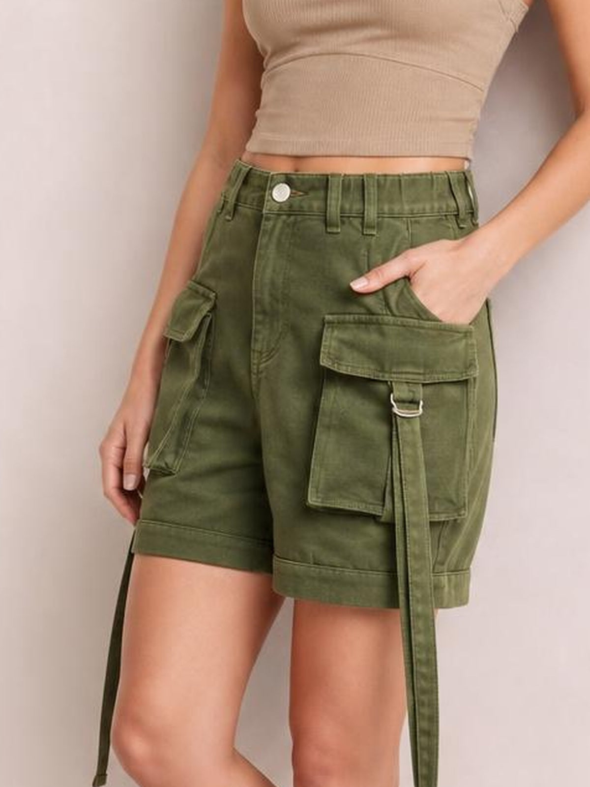 A model wears olive green high-waist denim cargo shorts with a relaxed silhouette, belt loops, button closure, and multiple flap pockets, one hand tucked inside.