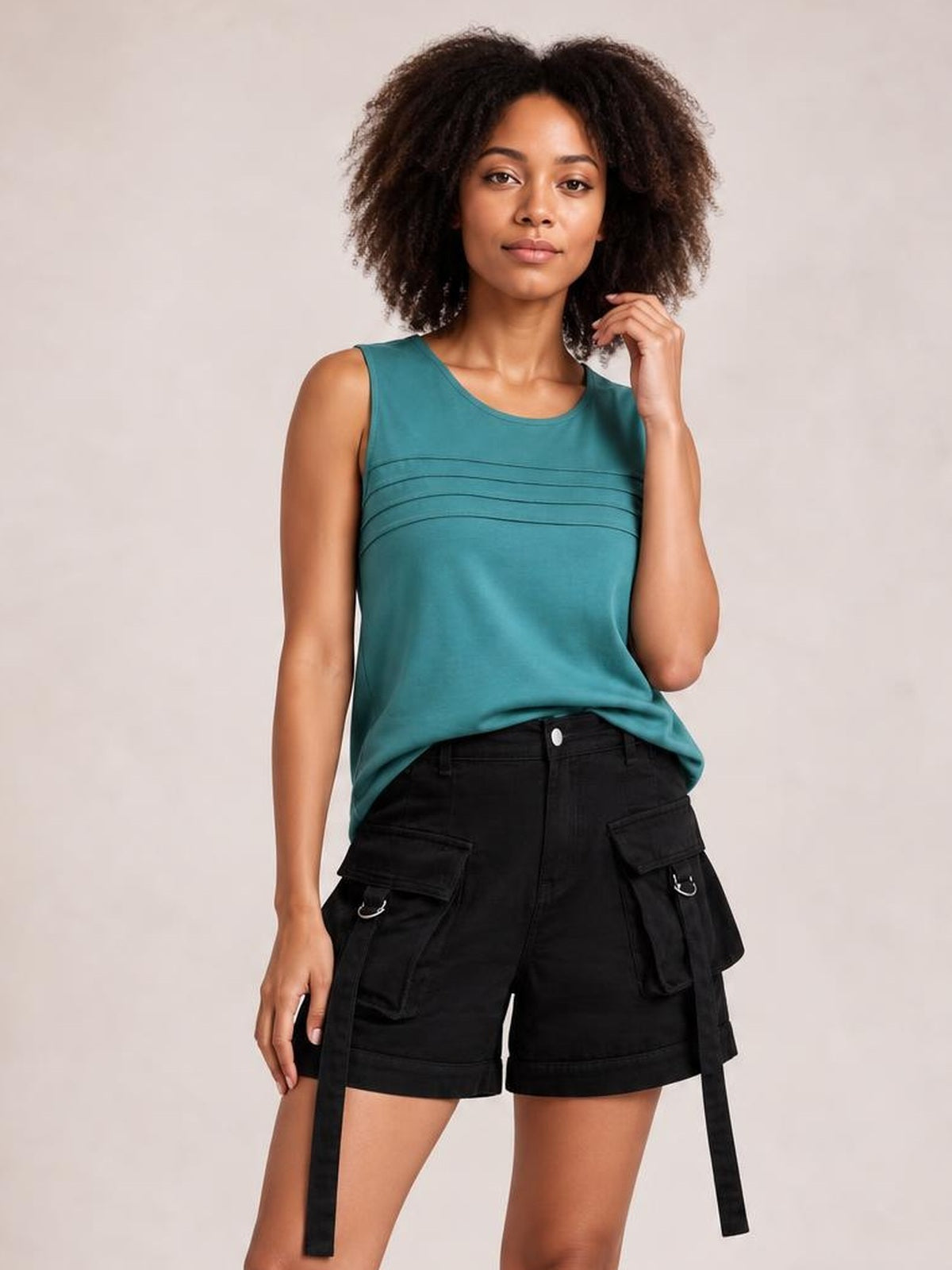 A woman modeling a sleeveless teal top with horizontal seam details and black cargo shorts, standing against a plain light beige studio background.