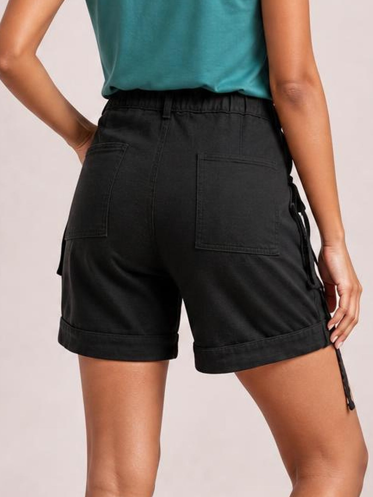 A model poses from the back in solid black high-waist denim cargo shorts with belt loops, large patch pockets, cuffed hems, and a relaxed fit.