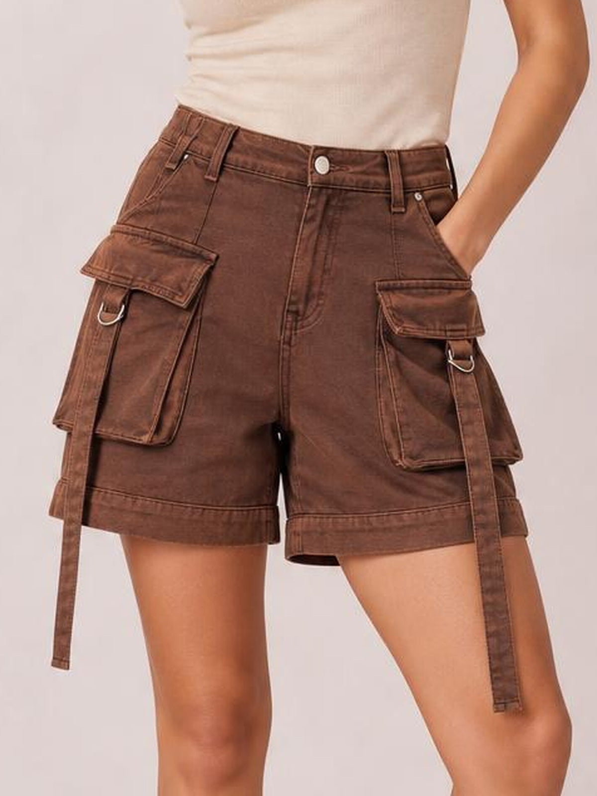 A model poses with one hand in the pocket, wearing brown high-waist denim cargo shorts with a relaxed fit, belt loops, flap pockets, and hanging straps.