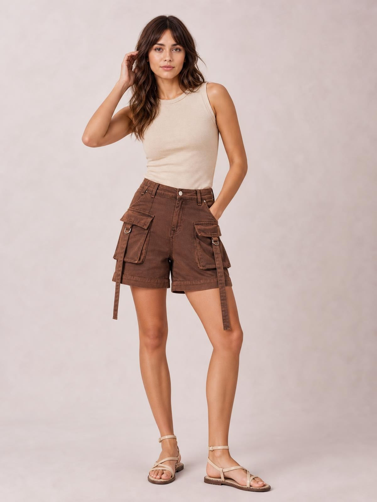 A model poses in solid brown high-waist denim cargo shorts with a structured silhouette, multiple flap pockets, belt loops, and hanging utility straps.
