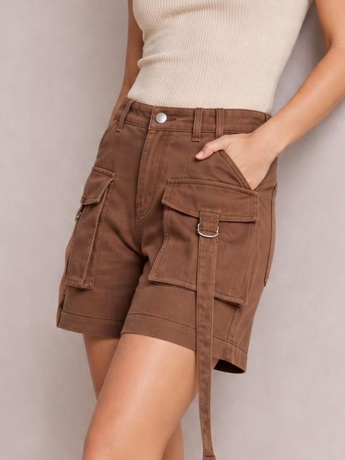 Model wears brown high-waist denim cargo shorts with a structured silhouette, belt loops, stitched panels, and multiple flap pockets while posing with one hand in a pocket.