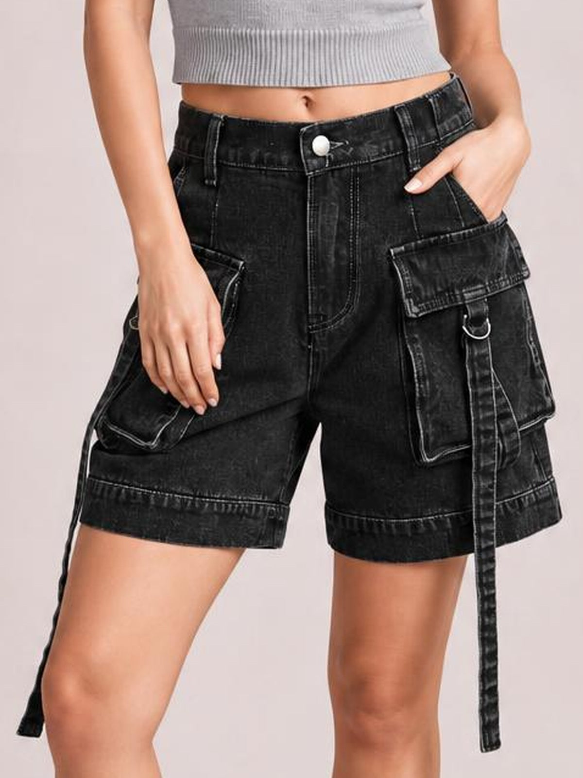 A model poses in black high-waist denim cargo shorts with a relaxed knee-length silhouette, multiple flap pockets, contrast stitching, and hanging utility straps.