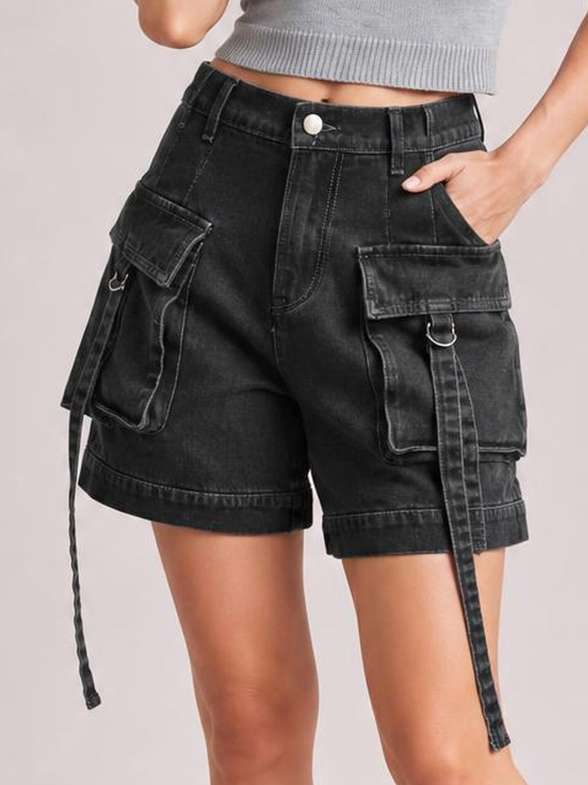 A model poses in black high-waist denim cargo shorts with a button fly, belt loops, relaxed silhouette, oversized flap pockets, straps, and metal rings.