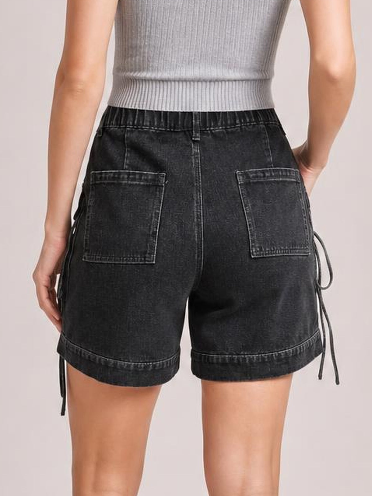 A model wears black high-waist denim cargo shorts with a relaxed silhouette, large patch pockets, stitched seams, rolled cuffs, and side tie details.