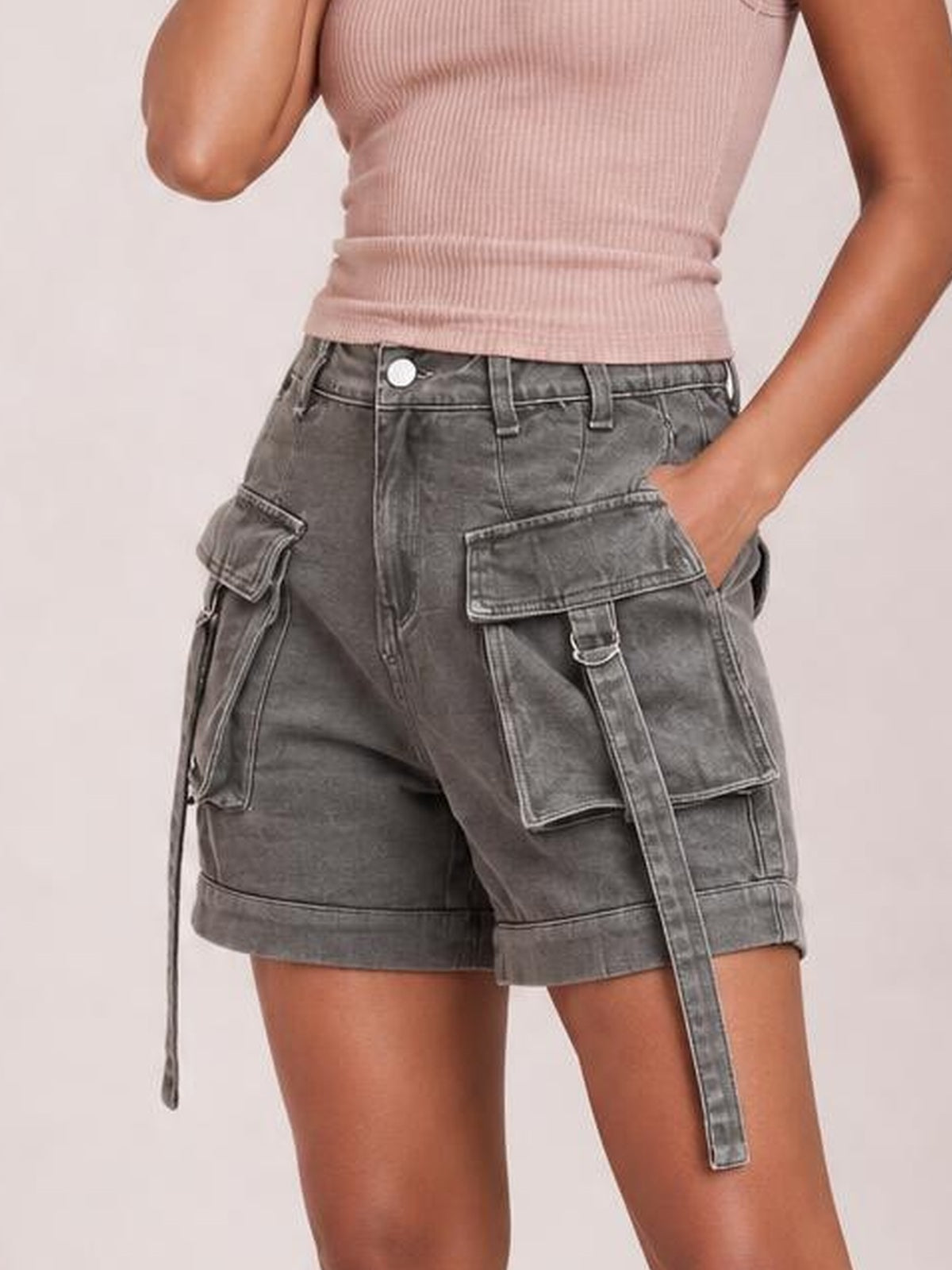 A model poses with one hand in the pocket, showcasing gray high-waist denim cargo shorts with multiple flap pockets, belt loops, stitched panels, and cuffed hems.