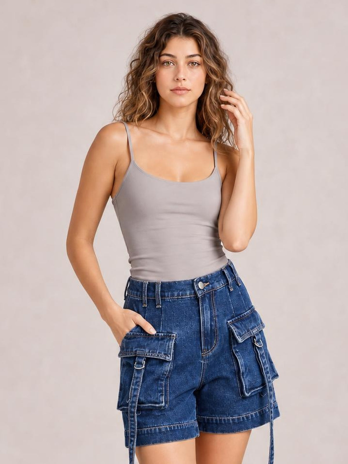 A woman modeling a light gray spaghetti-strap camisole top paired with high-waisted blue denim cargo shorts against a neutral beige studio background.