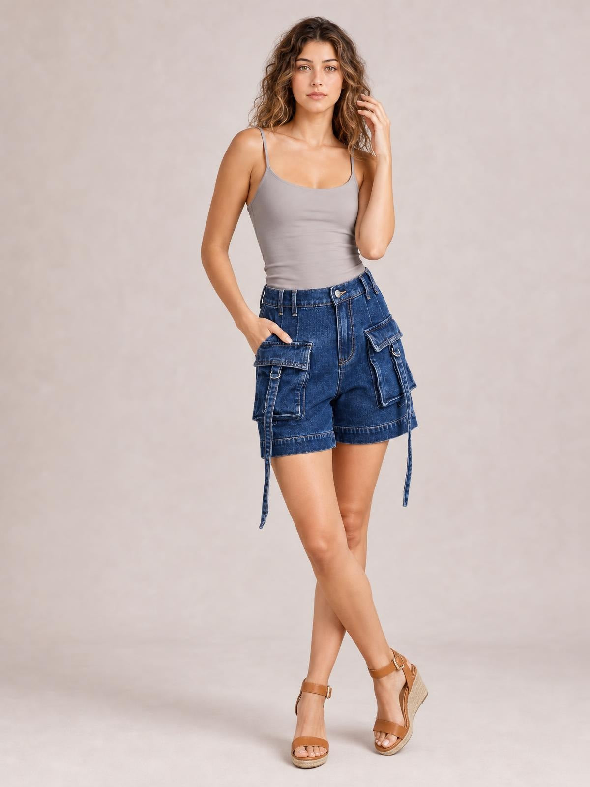 A model poses with one hand in the pocket, wearing blue high-waist denim cargo shorts with a relaxed fit, multiple flap pockets, and long hanging straps.