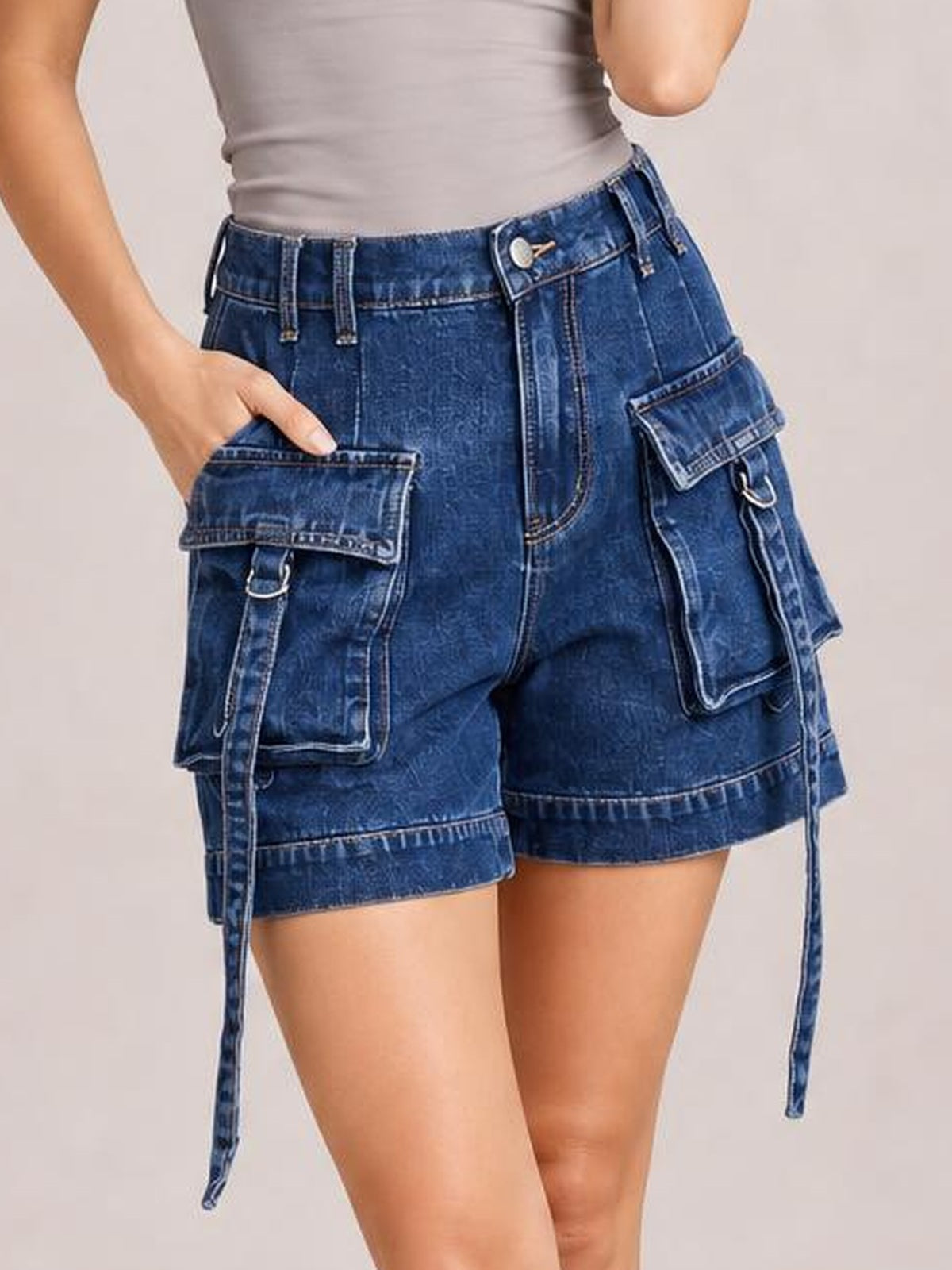 A model wears solid blue high-waist denim cargo shorts with a relaxed silhouette, multiple flap pockets, belt loops, and rolled hems, posing with one hand in a pocket.