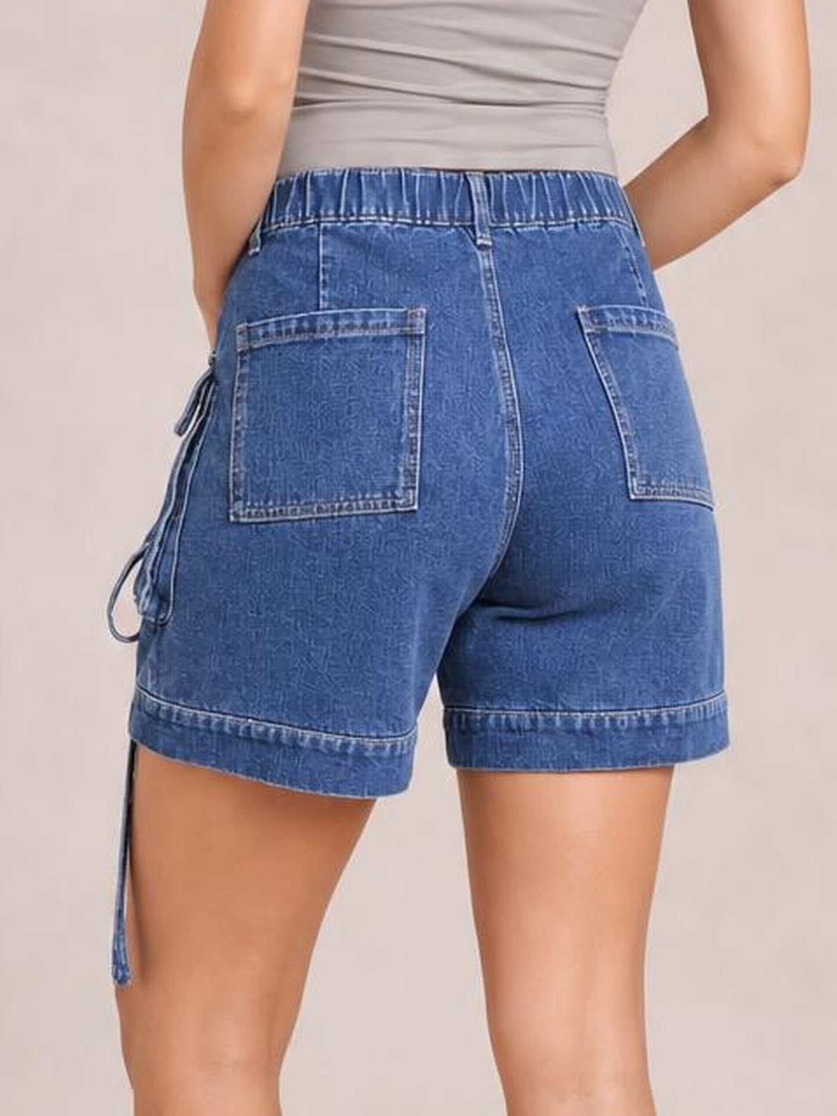 Model shown from behind wearing blue high-waist denim cargo shorts with a relaxed fit, multiple patch pockets, side drawstring detail, and rolled hems.