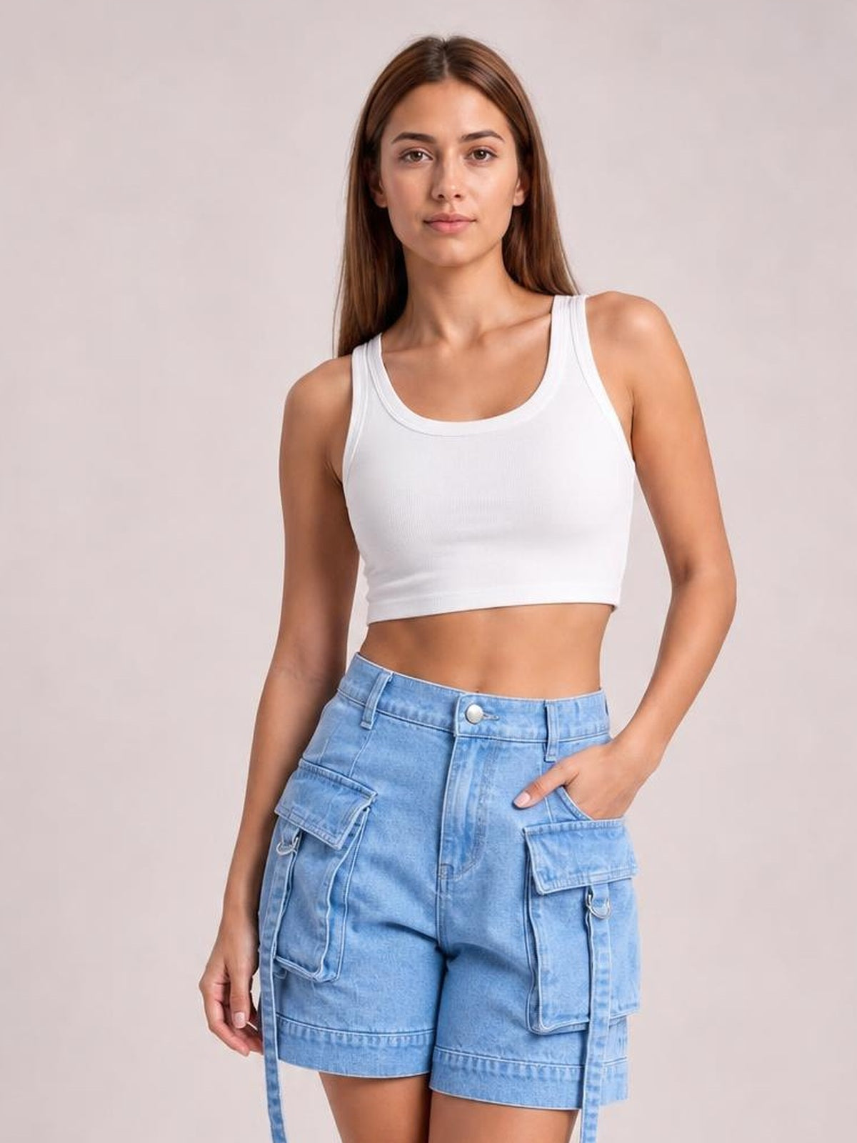 A woman modeling a white ribbed scoop-neck crop top and light blue denim cargo shorts against a solid light beige studio background.