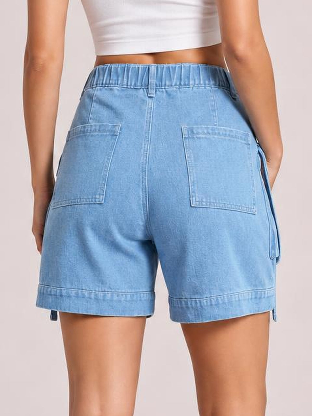 Model shown from behind wearing light blue high-waist denim cargo shorts with an elasticized waistband, roomy silhouette, large back patch pockets, and a side cargo pocket.