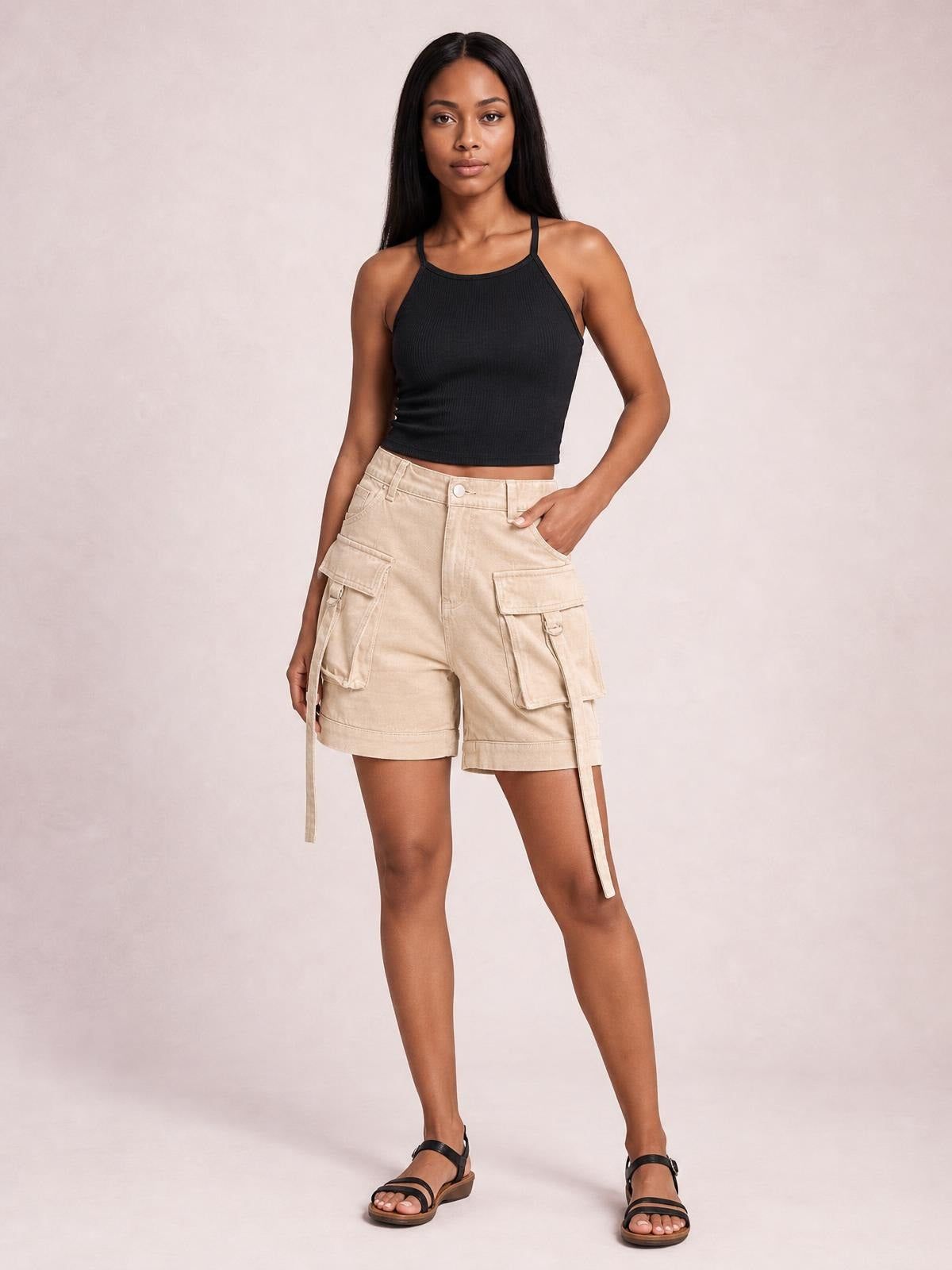 Model wears solid beige high-waist denim cargo shorts with a structured relaxed fit, button closure, rolled hems, and multiple flap pockets while standing with one hand in a pocket.