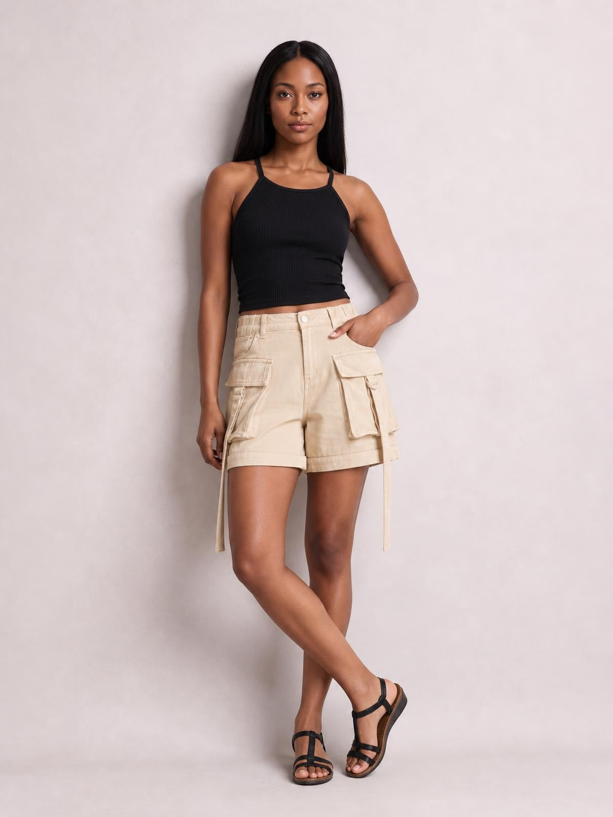 A model poses with one leg crossed in solid beige high-waist denim cargo shorts featuring a relaxed silhouette, cuffed hems, and multiple flap pockets.