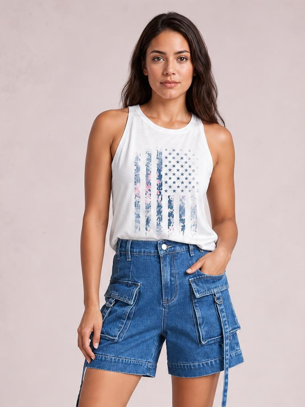 A woman modeling a white sleeveless tank top with a distressed blue American flag graphic, paired with blue denim cargo shorts, against a solid light pink background.