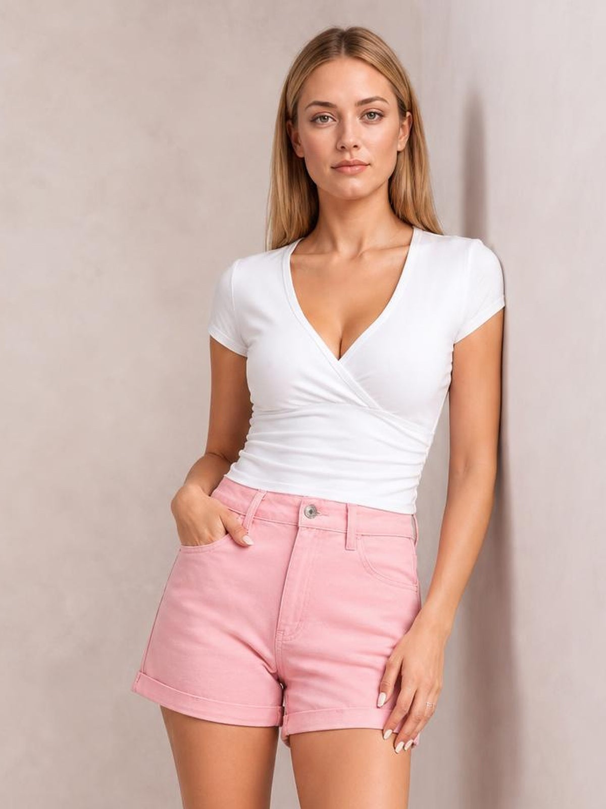 A woman modeling a white short-sleeve wrap-style V-neck top and light pink denim shorts against a neutral beige wall.