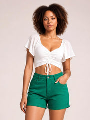 A woman modeling a white short-sleeve crop top with a ruched front and drawstring, paired with bright green shorts, against a solid light beige background.
