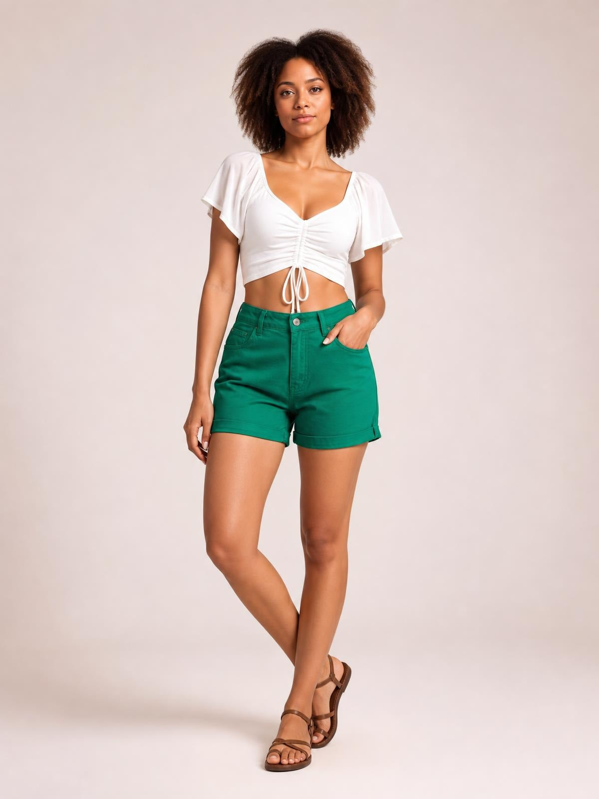 A model poses with crossed legs and one hand in the pocket, wearing green high-waist denim shorts with a button closure, pockets, and cuffed hems.