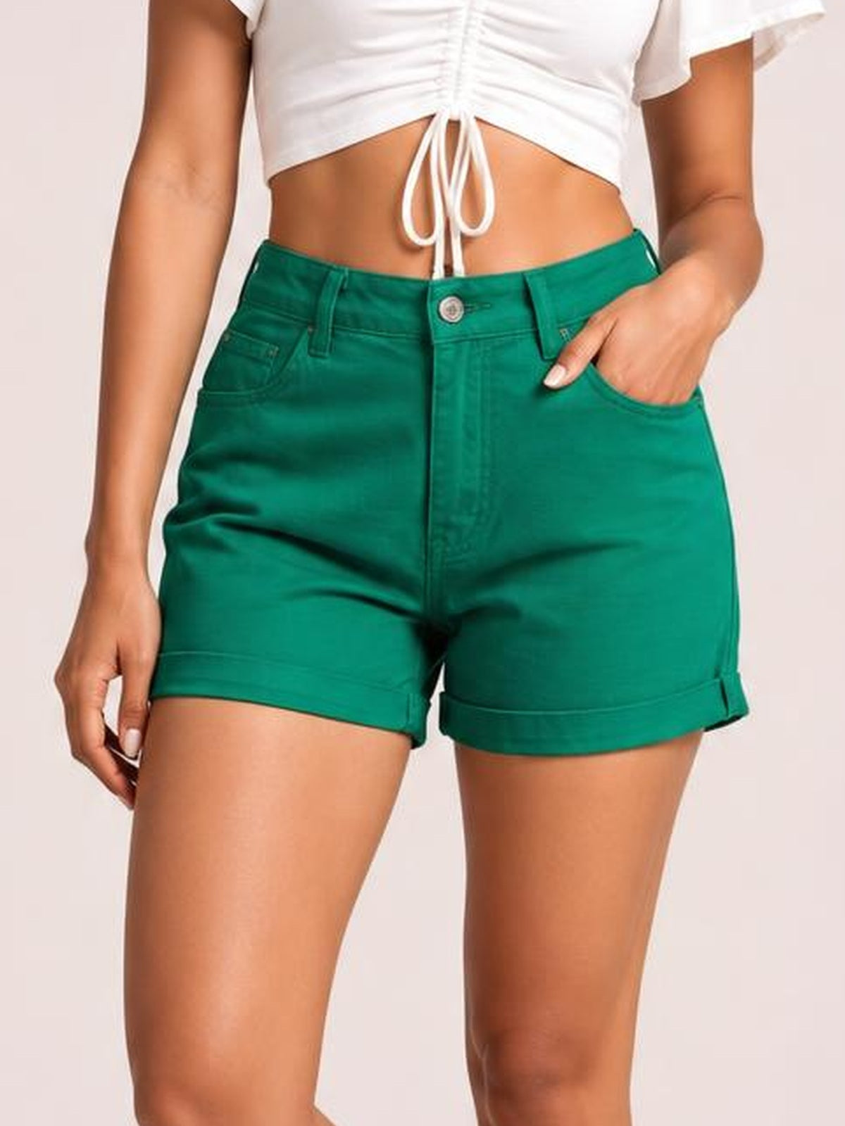 Model wears green high-waist denim shorts with a fitted silhouette, button closure, front pockets, belt loops, and neatly cuffed hems.