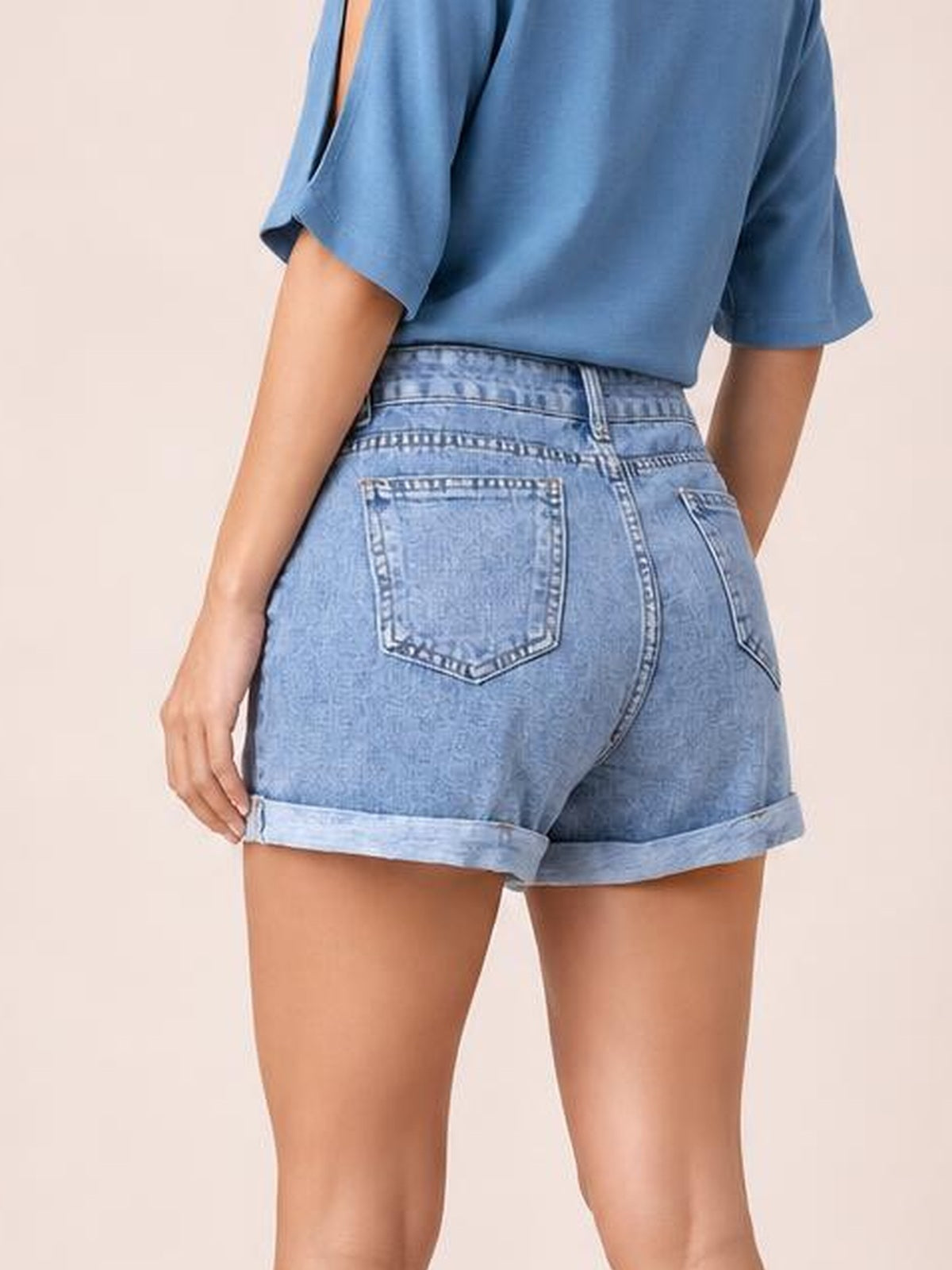 A model wears light blue high-waist denim shorts in a back view, featuring rolled cuffs, rear patch pockets, and a structured fitted silhouette.