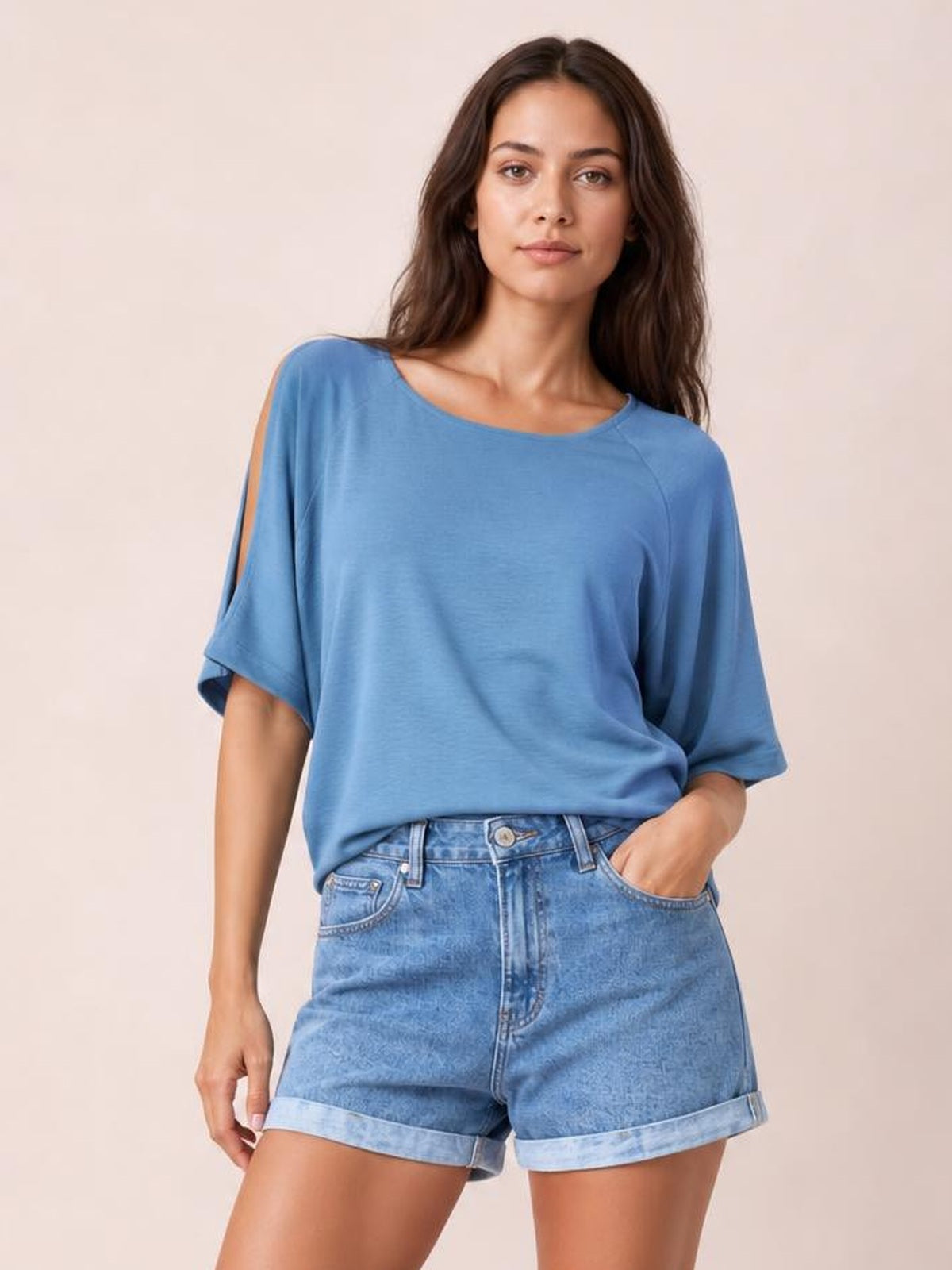 A woman modeling a light blue short-sleeve top with shoulder cutouts and light-wash denim shorts against a plain beige studio background.