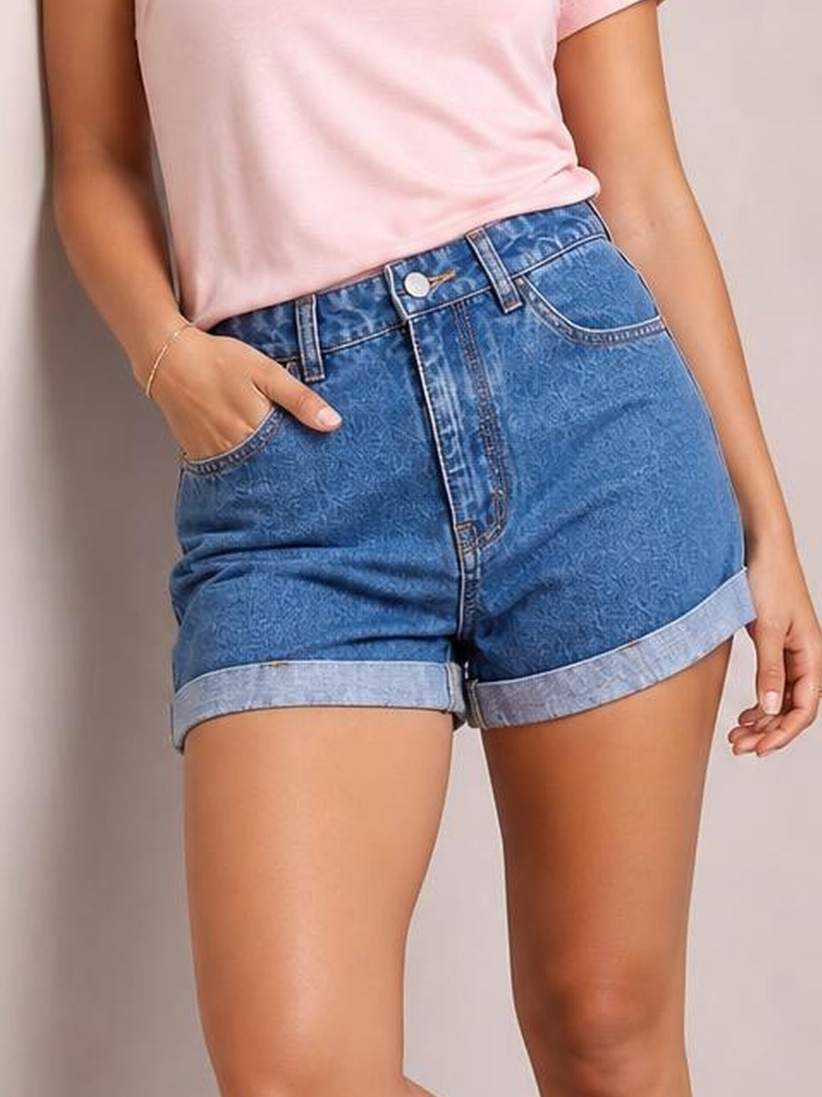 A model wears high-waist blue denim shorts with a button fly, front pockets, belt loops, and rolled cuffs, standing with one hand in a pocket.