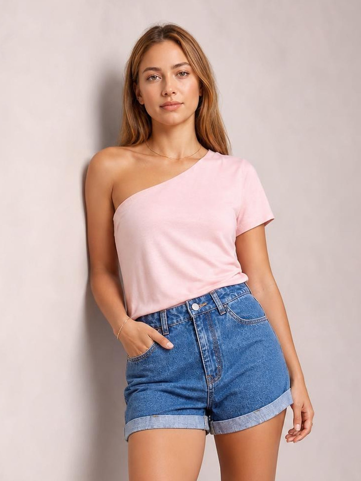 A woman modeling a light pink one-shoulder short-sleeve top paired with blue denim shorts, standing against a neutral light gray wall.