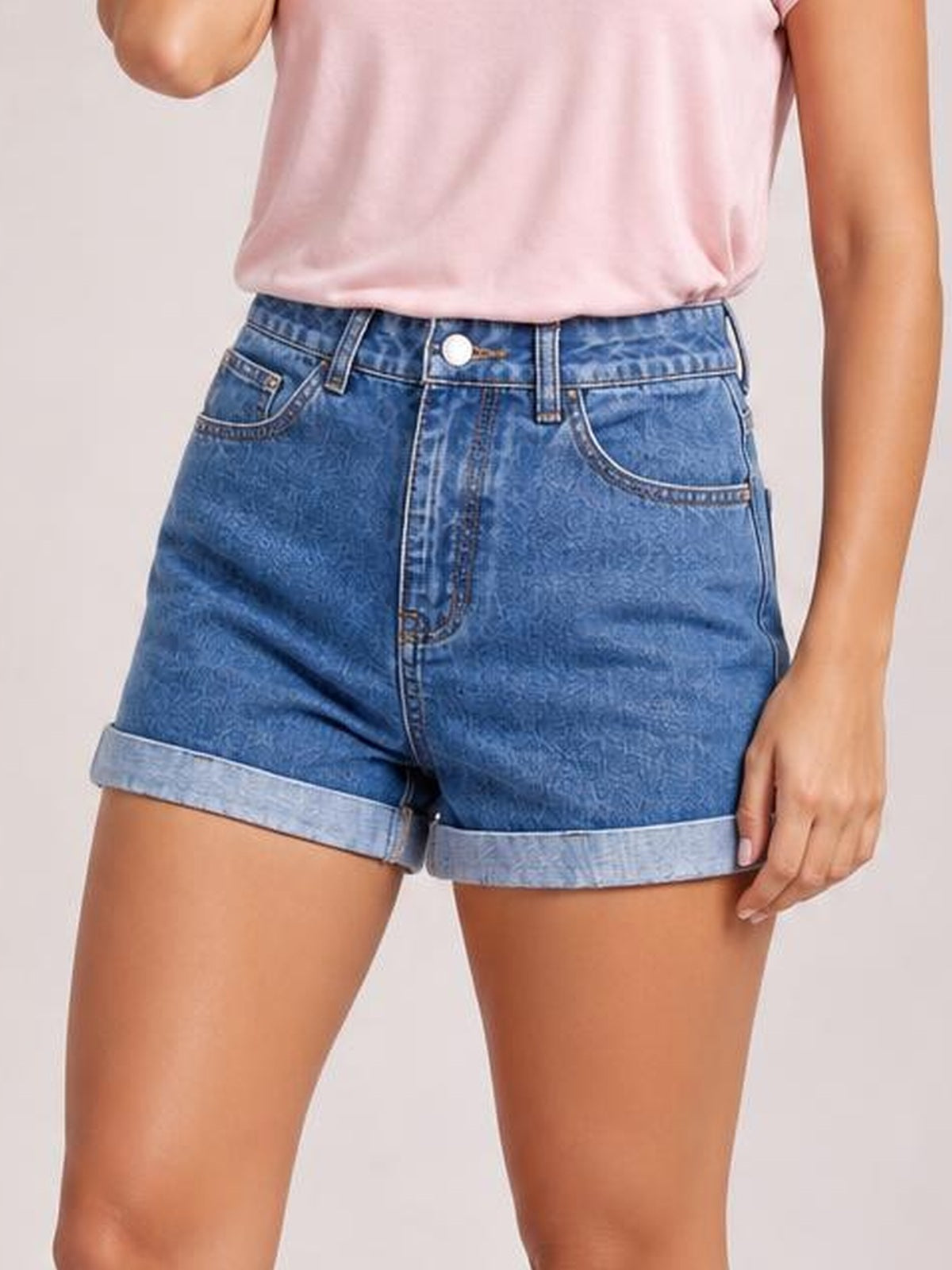 A model stands wearing blue high-waist denim shorts with a button closure, front pockets, structured silhouette, and rolled cuffs.