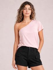 A woman modeling a light pink one-shoulder short-sleeve t-shirt paired with black cargo shorts, set against a solid pale pink studio background.
