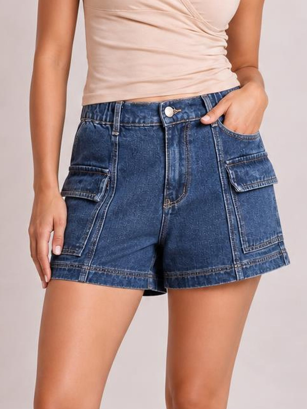 Model wearing high-waist dark blue denim shorts with a relaxed silhouette, button fly, contrast seam details, and multiple flap pockets, one hand tucked inside.