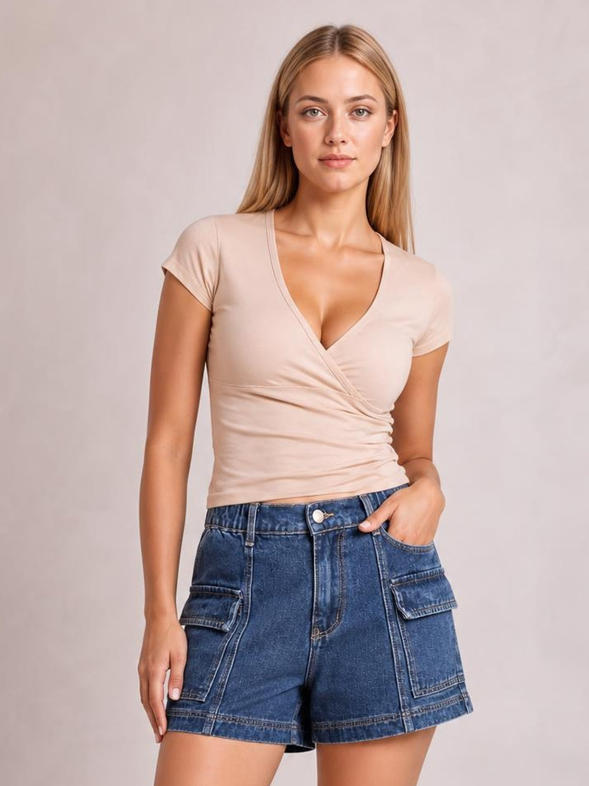 A woman modeling a beige short-sleeve wrap-style V-neck top paired with blue denim cargo shorts against a plain light gray studio background.