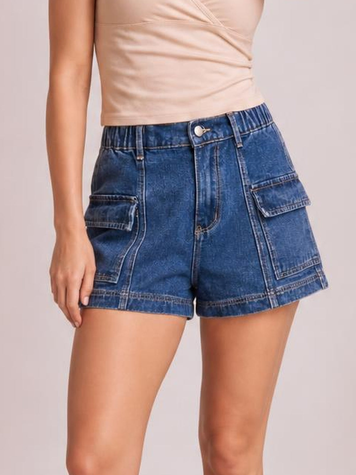 Model wears high-waist blue denim shorts with a relaxed silhouette, zip fly, belt loops, seam details, and large flap cargo pockets.