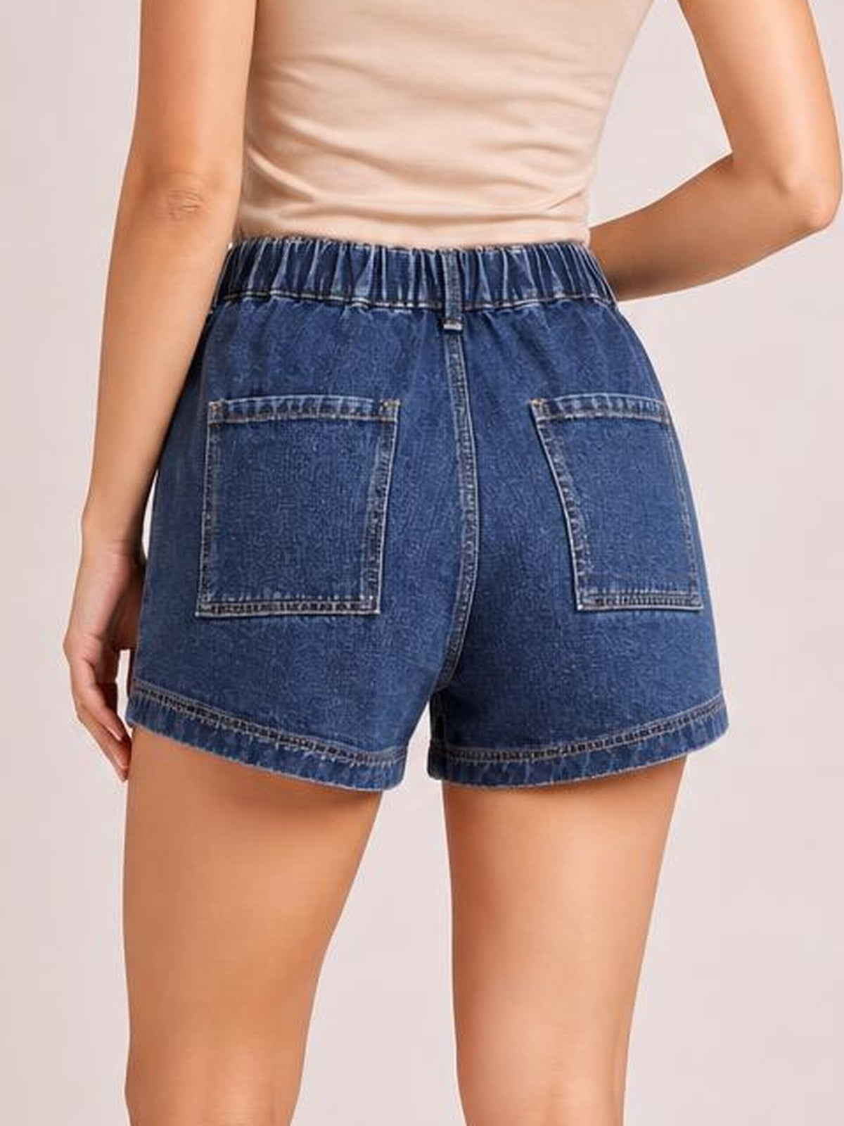 A model stands with one hand at her side and the other at her waist, showing blue high-waist denim shorts with an elastic waistband, seam details, patch pockets, and stitched hems.