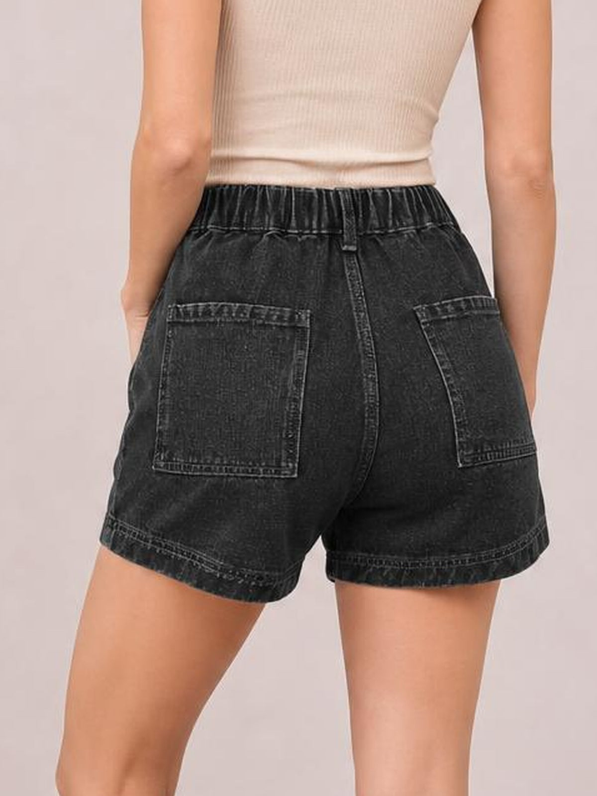 Model shown from behind wears black high-waist denim shorts with belt loops, contrast seam detailing, two back pockets, and a relaxed knee-length silhouette.