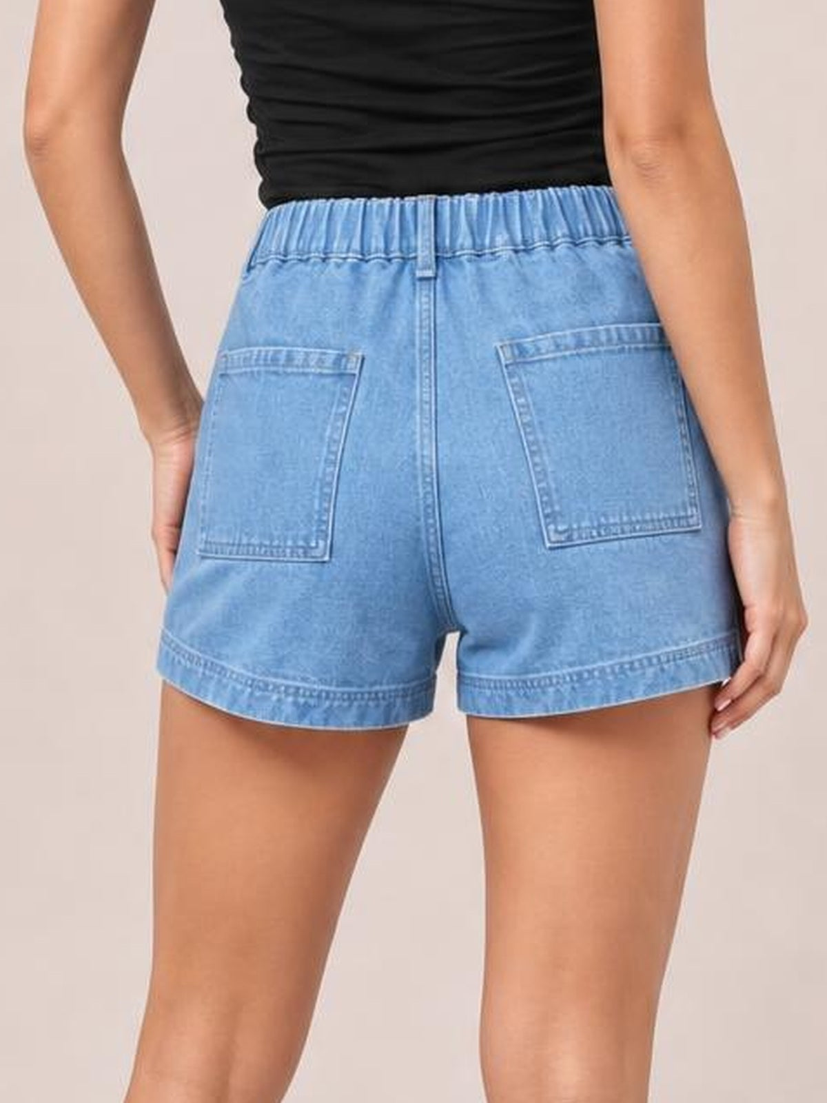 Model shown from behind wears light blue high waist denim shorts with an elastic waistband, stitched seam details, back patch pockets, and a relaxed short silhouette.