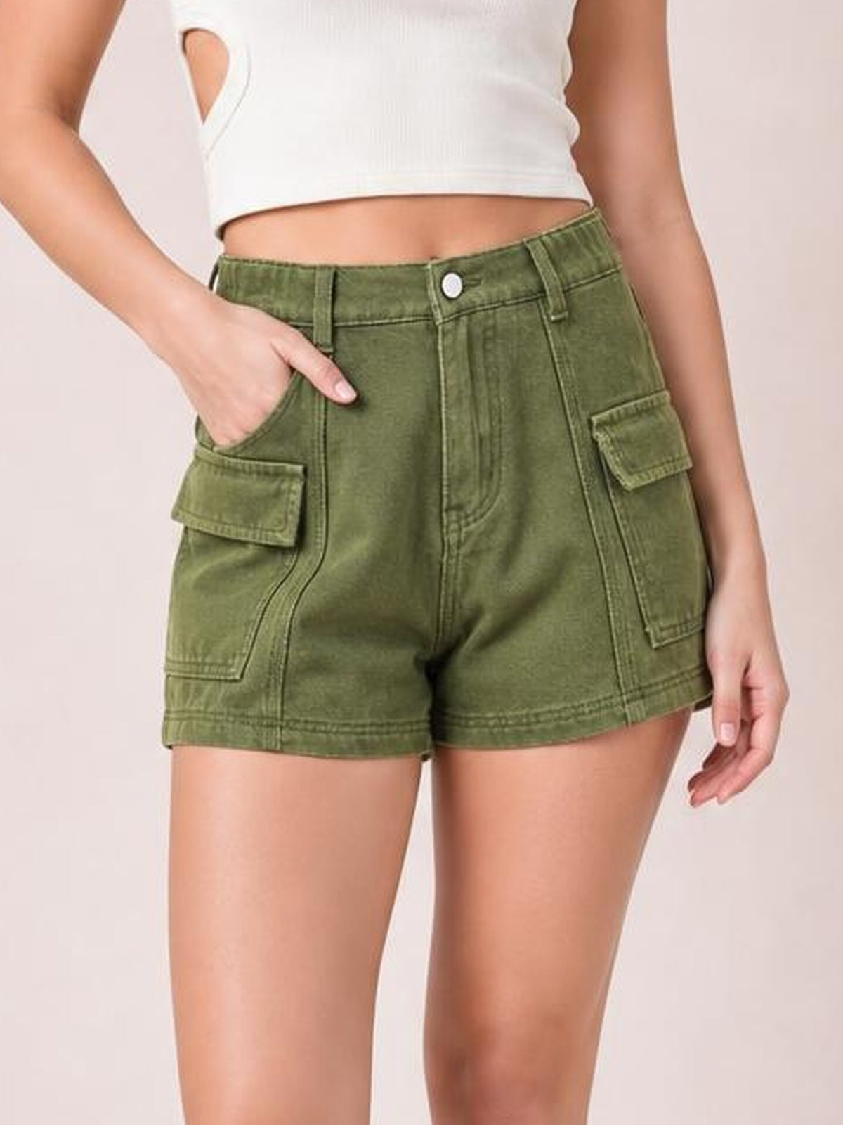 Model wears olive green high-waist cargo shorts with belt loops, button closure, seam detailing, flap pockets, and a relaxed straight-leg silhouette.